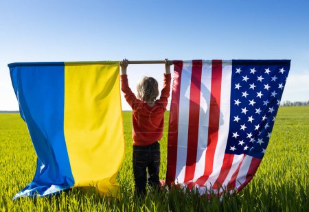 Photo for Child on bright sunny day stands back and holds two large flags - American and Ukrainian. concept of cooperation and friendship between two countries. US help to Ukraine to win war with Russia. - Royalty Free Image