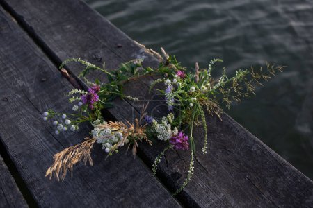 Téléchargez les photos : Couronne tissée de fleurs sauvages se trouve sur une jetée en bois près de l'eau. humeur romantique. Vacances pour les amoureux de la nature. Traditions pour les vacances d'Ivan Kupala. Profitez de l'été, détendez-vous - en image libre de droit