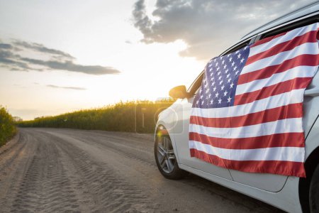 Photo for Large US flag is affixed to side of car driving down country road through rapeseed fields at sunset. USA Independence Day July 4th. Pride, freedom, patriotism, democracy. Travel around country - Royalty Free Image
