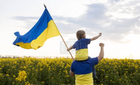 Foto de Padre con hijo sentado sobre hombros, en camisetas de color amarillo-azul con gran bandera de Ucrania en las manos contra el telón de fondo del campo de colza amarillo floreciente. Educación patriótica. Orgullo, libertad. Gloria a Ucrania - Imagen libre de derechos