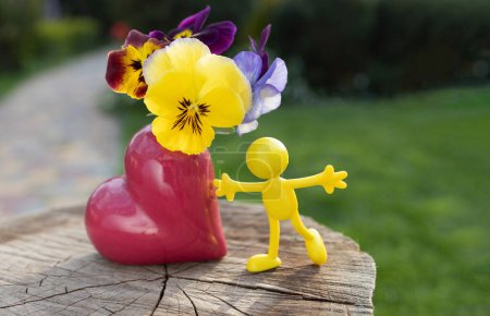 Foto de Figura miniatura de juguete brillante de un hombre amarillo cerca de un jarrón en forma de corazón con pantaletas. concepto de amor, alegría, felicidad. pensamiento positivo. regalo de flores. infancia divertida - Imagen libre de derechos