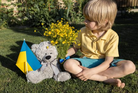 Photo for Teddy bear and a Ukrainian child in yellow and blue clothes are sitting on the grass. War and social problems in Ukraine. Independence Day in immigration. loneliness, nostalgia for the homeland - Royalty Free Image