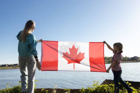 woman and a child, mother and son, hold the Canadian flag on a sunny day against the background of the sky and the river. Travel, immigration. Canada Day. Pride, freedom, national symbol