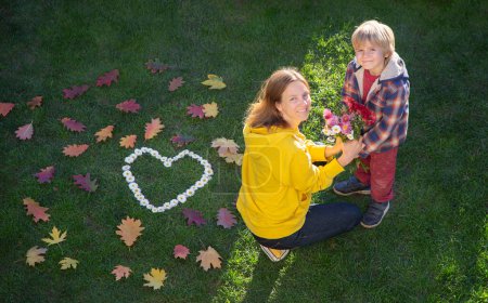 Téléchargez les photos : Jour d'automne. Son fils donne des fleurs à sa mère assise sur l'herbe. Les feuilles d'automne sont éparpillées autour et un cœur de chrysanthèmes est posé. Vacances en famille dans la nature. Relations tendres dans la famille - en image libre de droit