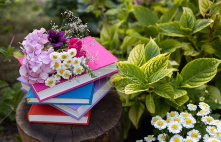 Téléchargez les photos : Pile de plusieurs livres multicolores se trouvent dans le jardin sur un moignon parmi les fleurs. Le concept de lecture, d'éducation, de sagesse et de connaissance. désintoxication numérique. jour du livre. Retour à l'école - en image libre de droit
