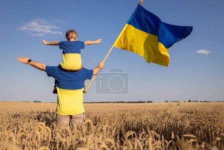 Foto de Ucranianos hombre con niño, padre con hijo, que se sienta sobre los hombros, de pie con la espalda con gran bandera de Ucrania entre de campo de trigo. Educación patriótica. Orgullo, fe en la victoria. Día de la Independencia. - Imagen libre de derechos