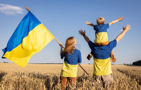 Téléchargez les photos : Heureuse famille ukrainienne en jaune-bleu t-shirts se tient parmi les épillets dans le champ de blé, enfant est assis sur les épaules de papa, femme tient drapeau de l'Ukraine. identité nationale. foi en la victoire. Jour de l'indépendance - en image libre de droit