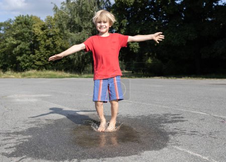 Téléchargez les photos : Pieds nus heureux enfant saute joyeusement dans une flaque transparente sur le trottoir après la pluie. Changements de temps en été. Intéressant amusant enfance. enfant positif, optimisme, bonne humeur - en image libre de droit