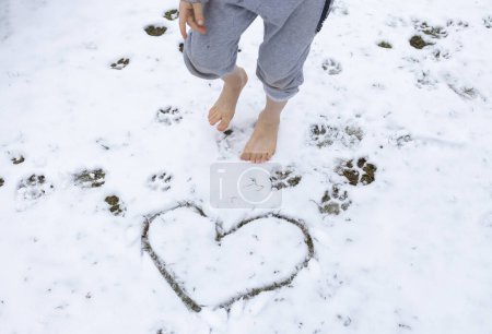 Téléchargez les photos : Enfant marche pieds nus sur la neige fraîchement tombée. Coeur dessiné et empreintes de pas d'un chien dans la neige. concept de soins de santé, durcissement, mode de vie sain. le froid. Amour hiver - en image libre de droit