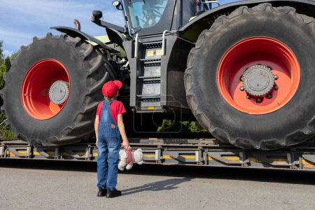 Child examines a huge tractor with large wheels loaded onto a truck for transportation. unrecognizable boy in denim overalls stands with his back in the frame.