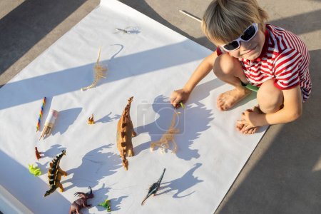 kleiner Junge spielt mit kontrastierenden Schatten von verschiedenen Figuren von Spielzeug-Dinosauriern. Kleiner Wissenschaftler, Ideen zur Entwicklung der Fantasie, Spiele für Kinder, Interesse an der Paläontologie