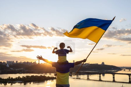 papa avec son fils, qui est assis sur les épaules, avec le grand drapeau de l'Ukraine sur le toit de la maison sur le remblai de la rivière Dniepr et le paysage de Kiev. L'éducation patriotique. foi en la victoire. arrêter la guerre. Jour de l'indépendance