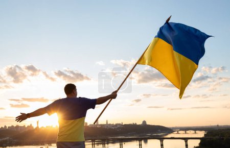 Foto de Hombre con gran bandera amarilla y azul ucraniana ondeando en la mano contra el cielo y el río Dniéper en Kiev al atardecer. Apoya a Ucrania. Día de la Independencia Fe en la victoria. detener la guerra. Soy ucraniano. Vista trasera - Imagen libre de derechos
