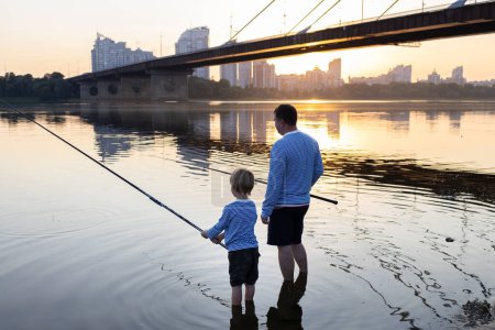 Téléchargez les photos : Père et fils pêchent ensemble debout avec des cannes à pêche dans l'eau de la rivière au coucher du soleil. le concept de la fête des pères. passe-temps amusant en famille pour le week-end. Hobby pour enfant et parent - en image libre de droit
