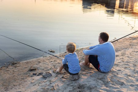 Téléchargez les photos : Papa et son fils se détendent ensemble, assis sur la rive de la rivière avec canne à pêche. le concept de la fête des pères. Amusement en famille pour le week-end. passe-temps commun pour le parent et la progéniture. être comme papa, éduquer la patience - en image libre de droit
