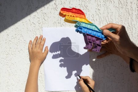 Kind zeichnet an einem sonnigen Tag einen kontrastierenden Schatten eines Dinosauriers auf Papier nach. glückliche, fröhliche Kindheit, Spielideen, Lebensstil, Entwicklung der Fantasie, kreative Spiele