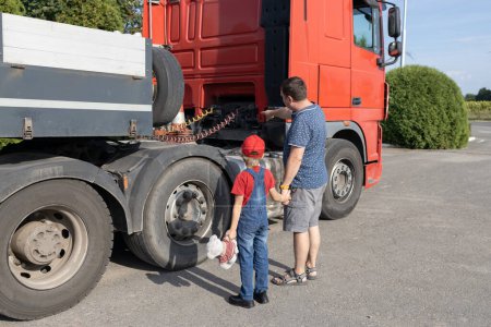 Téléchargez les photos : Homme et un garçon, père et fils, se tiennent devant un grand camion, examinant son mécanisme. La passion d'un enfant pour les voitures. Loisirs des hommes - en image libre de droit