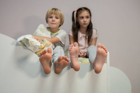 Two barefoot children are sitting next to each other on the bed. Early morning sleepy brother and sister, unwillingness to wake up. Selective focus on boy's bare feet