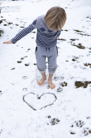 Téléchargez les photos : Enfant marche pieds nus sur la neige fraîchement tombée en hiver. Un cœur est tiré sur la neige. concept de soins de santé, durcissement, mode de vie sain, auto-défi. le froid. Amour hiver - en image libre de droit