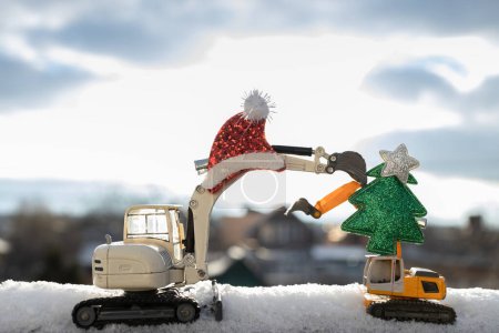 Foto de Saludos de Año Nuevo de negocios. excavadoras de juguete, árbol de Navidad, sombrero de Santa en la nieve contra el cielo. humor de vacaciones de invierno. Es hora de cambiar. tarjeta del humor de vacaciones de invierno - Imagen libre de derechos
