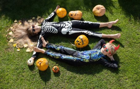 Foto de Dos niños. un niño y una niña en disfraces de Halloween yacen en la hierba con calabazas pintadas de naranja. Feliz infancia alegre. Hijos, amigos en una fiesta de vacaciones. Vista desde arriba - Imagen libre de derechos