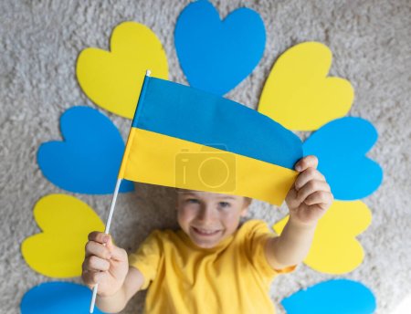 The child's hands hold the Ukrainian flag. the child lies among blue and yellow hearts. Independence Day. Love Ukraine, patriotism, peace and the anti-war concept. Top view, selective focus