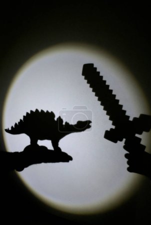 Foto de Interesante contraste de sombras en la pared de un dinosaurio de juguete y una espada. Juegos con luz y sombra. feliz infancia alegre, ideas para la tarde tiempo de la familia, estilo de vida, desarrollo de la imaginación - Imagen libre de derechos