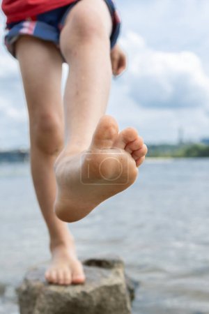 Téléchargez les photos : Pieds nus enfant lève le pied nu tout en se tenant sur une jambe sur une pierre dans la rivière. Vacances d'été et mode de vie sain concept. J'adore l'été, la détente, le soleil. C'est amusant d'aller de l'avant. vue du bas - en image libre de droit