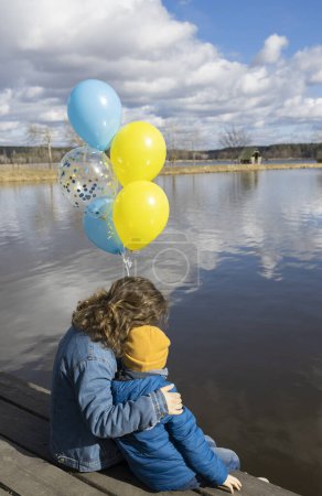 Téléchargez les photos : Des civils, des Ukrainiens, des réfugiés, un petit garçon méconnaissable et une jeune femme avec des ballons ukrainiens bleus et jaunes s'étreignent et s'assoient près du lac. Famille, unité. Arrêtez la guerre. soutenir l'Ukraine - en image libre de droit