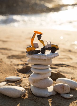 dos mini máquinas de construcción de juguetes naranja, una excavadora y una cargadora, se paran en una torre de piedras planas en la playa. Vacaciones en una empresa de construcción. Concepto de estabilidad en el negocio de la construcción