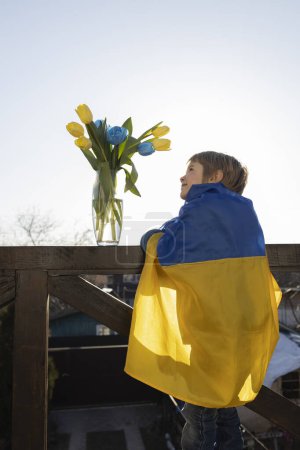 boy stands on the balcony wrapped in a Ukrainian flag admiring a bouquet of blue and yellow tulips. I'm proud to be Ukrainian. stand with Ukraine. Refugees, nostalgia. Love homeland. Stop the war