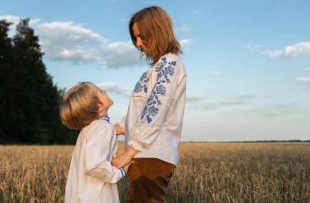 mom and son in embroidered national clothes stand among wheat field holding hands. Ukrainian friendly family, refugees, concept of unity, support, tenderness of motherhood, care. Stop war in Ukraine