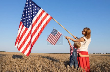 Große und kleine amerikanische Flagge in den Händen einer Frau und eines Kindes, die in einem vom Sonnenlicht erleuchteten Weizenfeld stehen. Unabhängigkeitstag der Vereinigten Staaten von Amerika. Stolz, Freiheit, Nationalsymbol