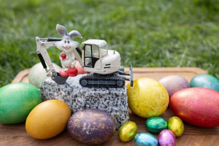 model of a toy excavator on a piece of granite, a souvenir rabbit, colorful eggs. Easter spring holiday concept for congratulations from construction companies