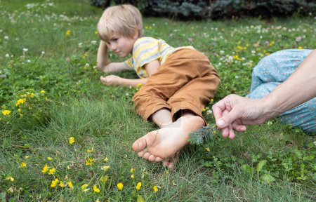 friendly cheerful family, mother plays with barefoot son lying on grass, tickling feet with fir twig. People having fun in summer park. positive thinking, good mood, lifestyle, joy of communication