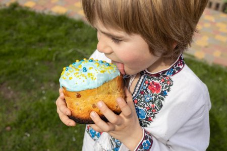 Junge schmeckt das süße Sahnehäubchen aus der Paska. Ein ukrainisches Kind im bestickten Kleid hält einen Osterkuchen in der Hand und leckt ihn mit der Zunge ab. Osterkonzept
