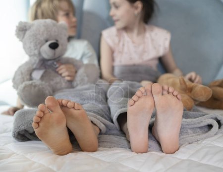 Foto de Dos niños descalzos, hermano y hermana, sentados en la cama, sosteniendo juguetes suaves en sus manos, se están divirtiendo comunicándose. niños de 7 a 8 años en casa en una cama grande, descanso matutino, alegría familiar - Imagen libre de derechos