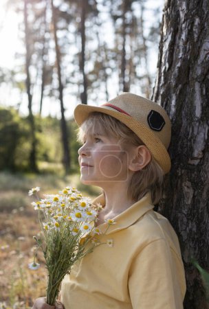 mignon garçon 7 ans dans la forêt près d'un arbre avec un bouquet de marguerites recueillies. atmosphère positive, enfance. cadeau pour la fête des mères. focus sélectif, promenades nature