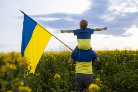 Vater und Sohn sitzen auf Schultern, die Arme ausgestreckt, stehen mit der Flagge der Ukraine inmitten blühender Rapsfelder. Bekleidet mit denselben gelben und blauen T-Shirts. Patriotismus, Stolz, Einheit, Independence Day