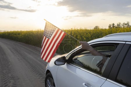 white car with an American flag hanging from the window drives along a country road among a blooming rapeseed field towards the setting sun . Travelling by car. Weekend trip, Bright future