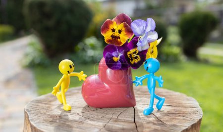 Figuras humanas de juguete miniatura en amarillo y azul cerca de un jarrón en forma de corazón con un ramo de maricones en un día soleado. pareja en el amor y el concepto de romance, amor y sentimientos tiernos