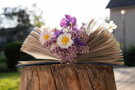 Auf Baumstumpf liegt ein aufgeschlagenes Buch, zwischen dessen Seiten ein Strauß verschiedener Blumen aus dem Garten liegt. Leseverständnis, Ästhetik, Weisheit und Wissen. Digitale Entgiftung. Buchtag. Grußkarte