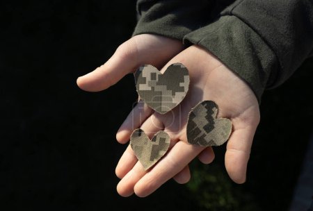 Drei aus Tarnstoff ausgeschnittene Herzen liegen auf der von der Sonne erleuchteten Kinderhandfläche. Schluss mit dem Krieg. Mit Liebe und Dankbarkeit gegenüber den Soldaten der Streitkräfte. Erfahrungen der Kinder mit Feindseligkeiten