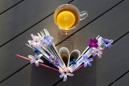 Tasse Tee auf Tisch, Buch und kleine Frühlingsblumen zwischen den Seiten. Bild eines friedlichen Morgens. Ruhe und Tee. Lesen Sie das Buch mit Vergnügen. Gemütliche heimelige Atmosphäre. Weisheit und Wissenskonzept, Draufsicht
