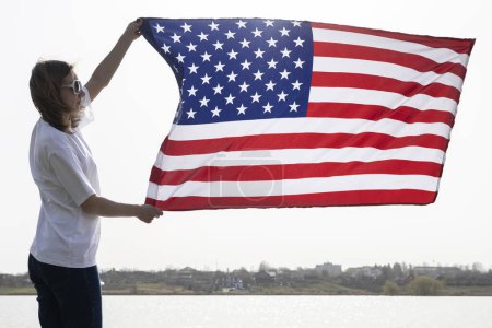 Photo for Woman stands near a lake and holds a large waving American flag in her hands. Independence Day. National symbol of freedom of the States of America. Bright future. patriotic holiday - Royalty Free Image