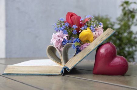 An open book lies on table, there are many different spring flowers on it, a heart made of pages. Read the book with pleasure. Concept of wisdom and knowledge, aesthetics, education. Digital detox