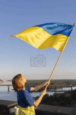 woman holds a large yellow-blue Ukrainian flag fluttering in the wind while standing high on the roof in Kyiv. Drawing attention to military actions in Ukraine. Be proud to be Ukrainian. stop the war