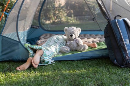bare feet of a child sleeping lying on his stomach in a tent with his favorite teddy bear. Healthy lifestyle, little tourist, family vacation, summer fun. Interesting childhood, tourism