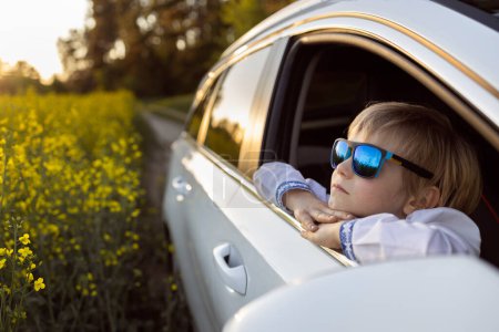 Das glückliche Gesicht eines kleinen Jungen mit blauer Sonnenbrille blickt aus dem offenen Autofenster und schaut sich interessiert um. Im Sommer mit der Familie mit dem Auto unterwegs