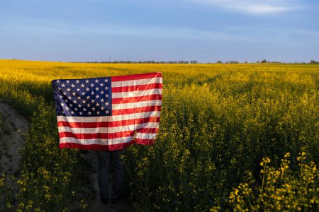 Große amerikanische Flagge inmitten eines blühenden gelben Rapsfeldes und des Himmels. Independence Day. Nationales Symbol der Freiheit der Staaten von Amerika